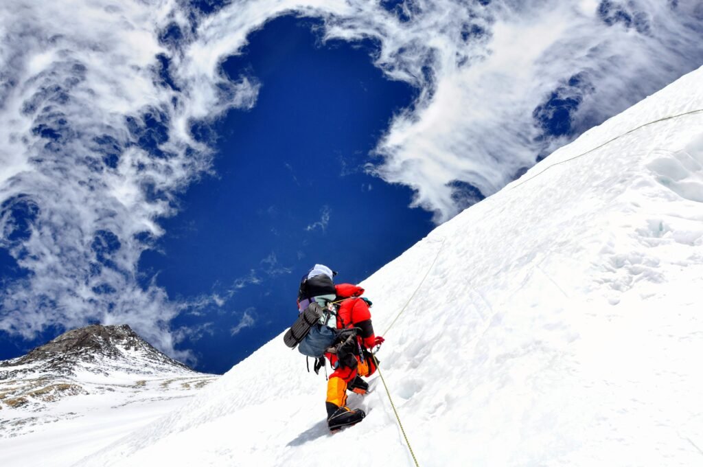 Climber in red ascends snowy mountain with blue sky and wispy clouds overhead.